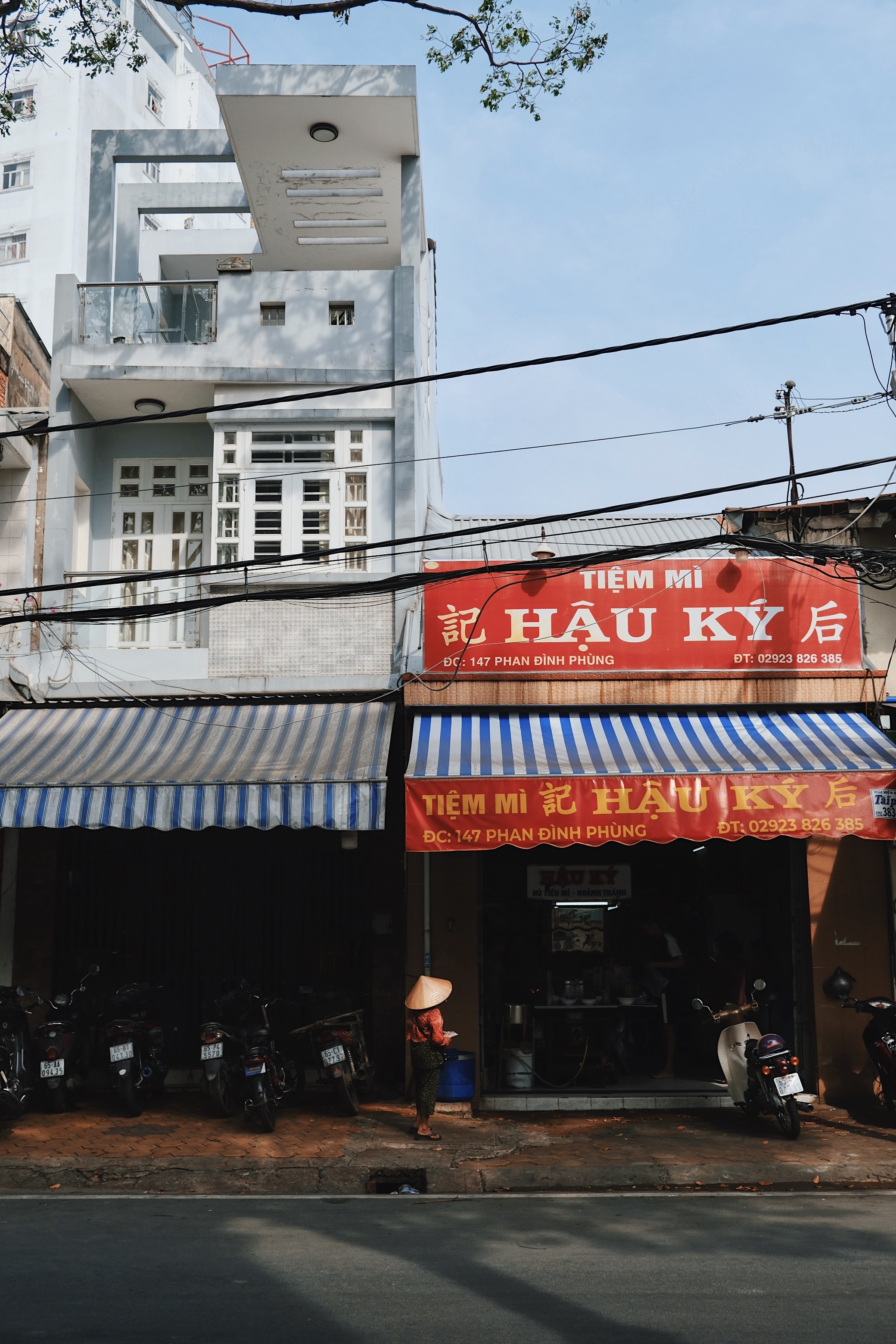 A Vietnamese woman wearing a traditional conical hat walks past a modernist building in Cần Thơ, illustrating the fusion of traditional daily life and contemporary architecture.