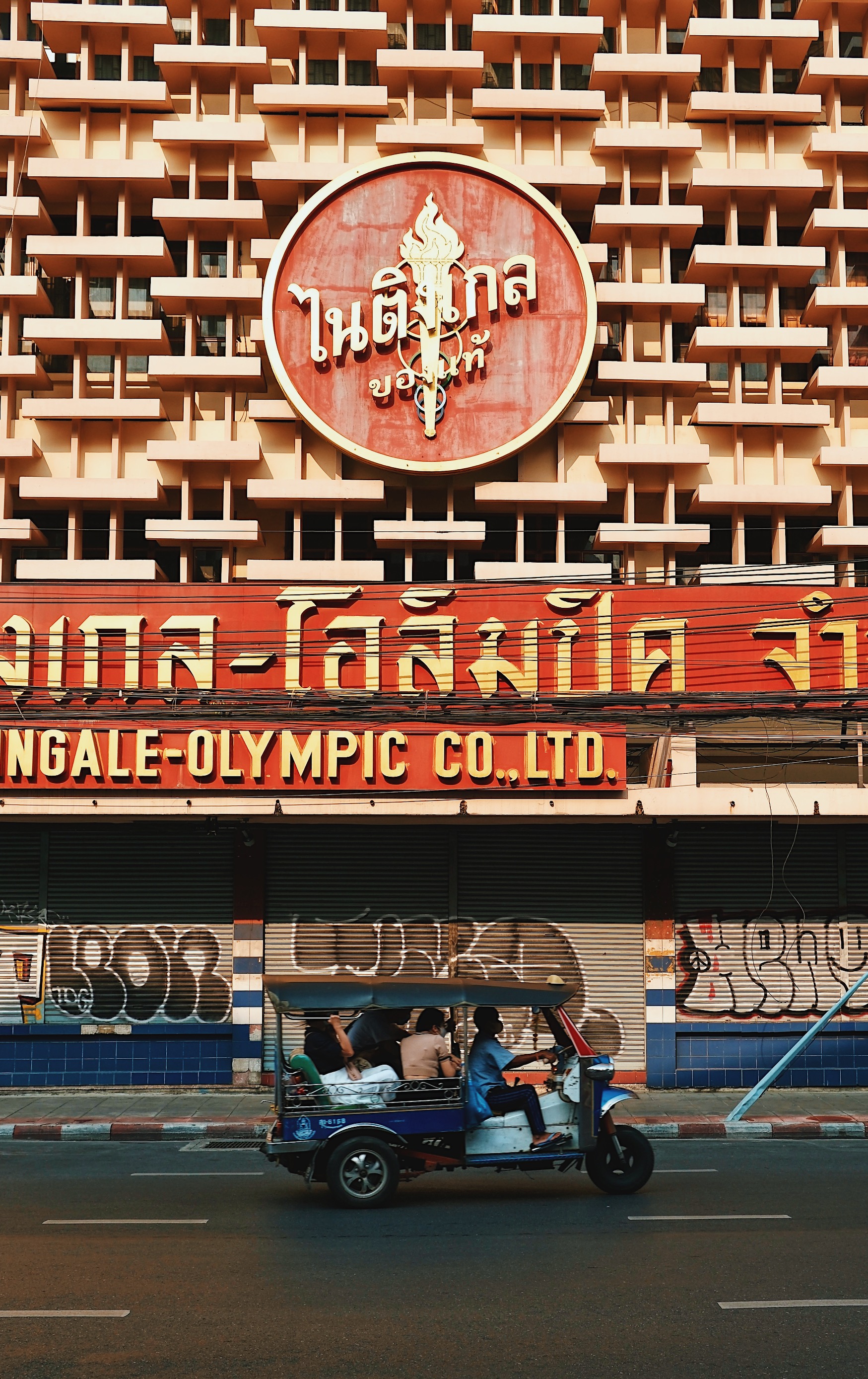 A traditional Thai tuk-tuk speeds past a weathered modernist concrete building in Bangkok, showcasing the city’s mix of old and new.