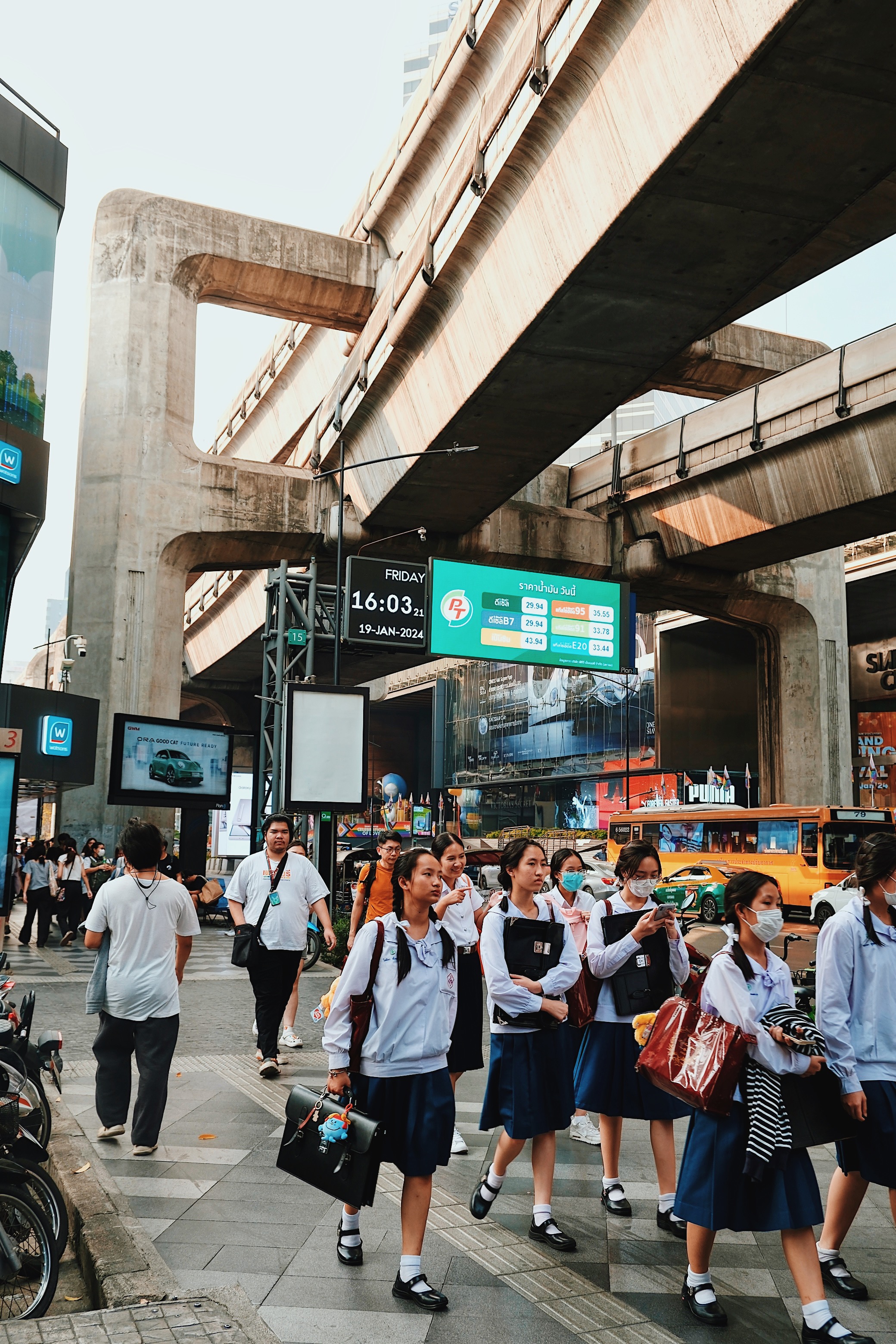  Students in uniforms walk through Siam Square, Bangkok, beneath the city's elevated Skytrain tracks, surrounded by modernist concrete structures.
