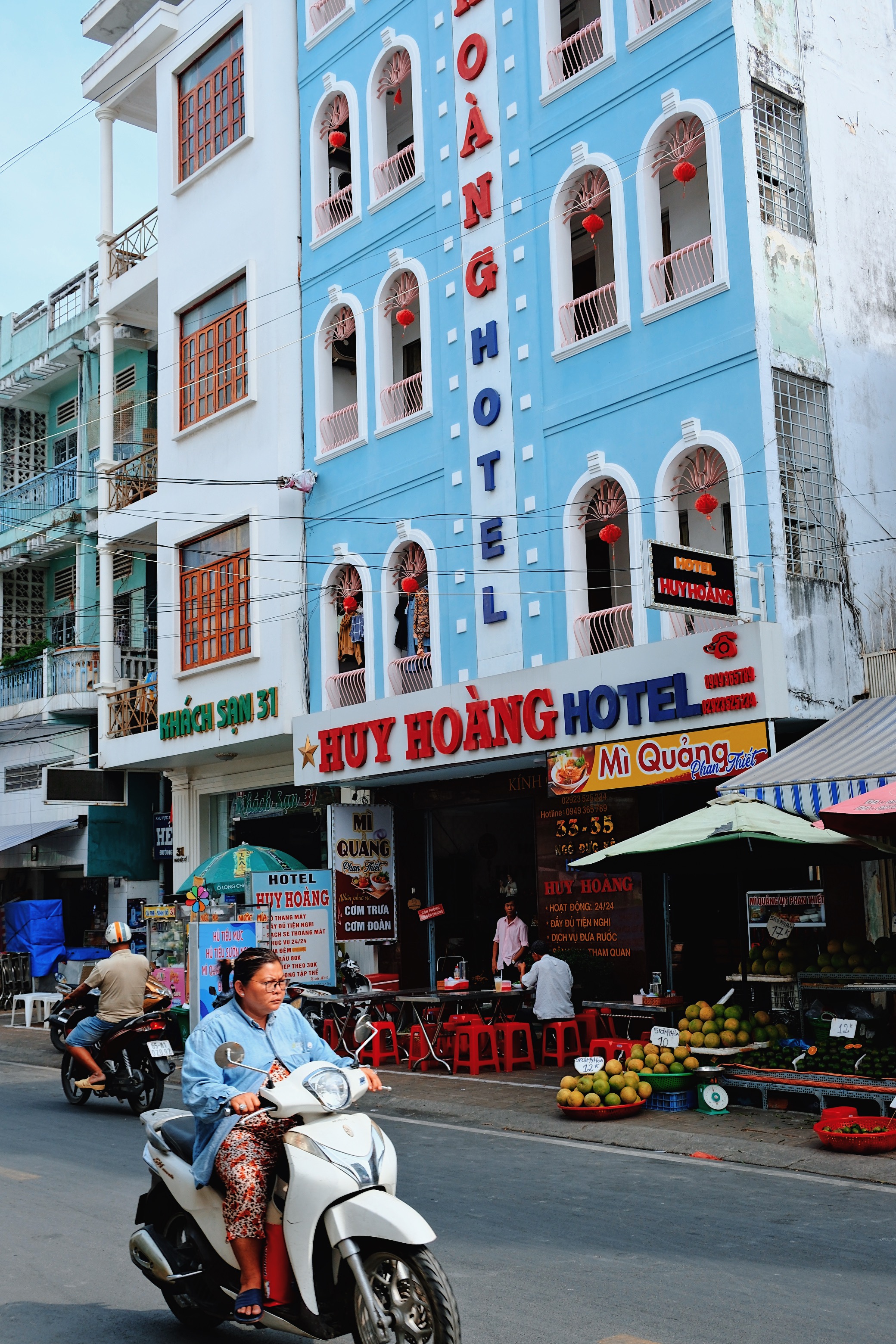 A pastel blue hotel in Can Tho, Vietnam with arched windows and red lanterns, as a woman in a denim jacket rides her scooter past fruit vendors