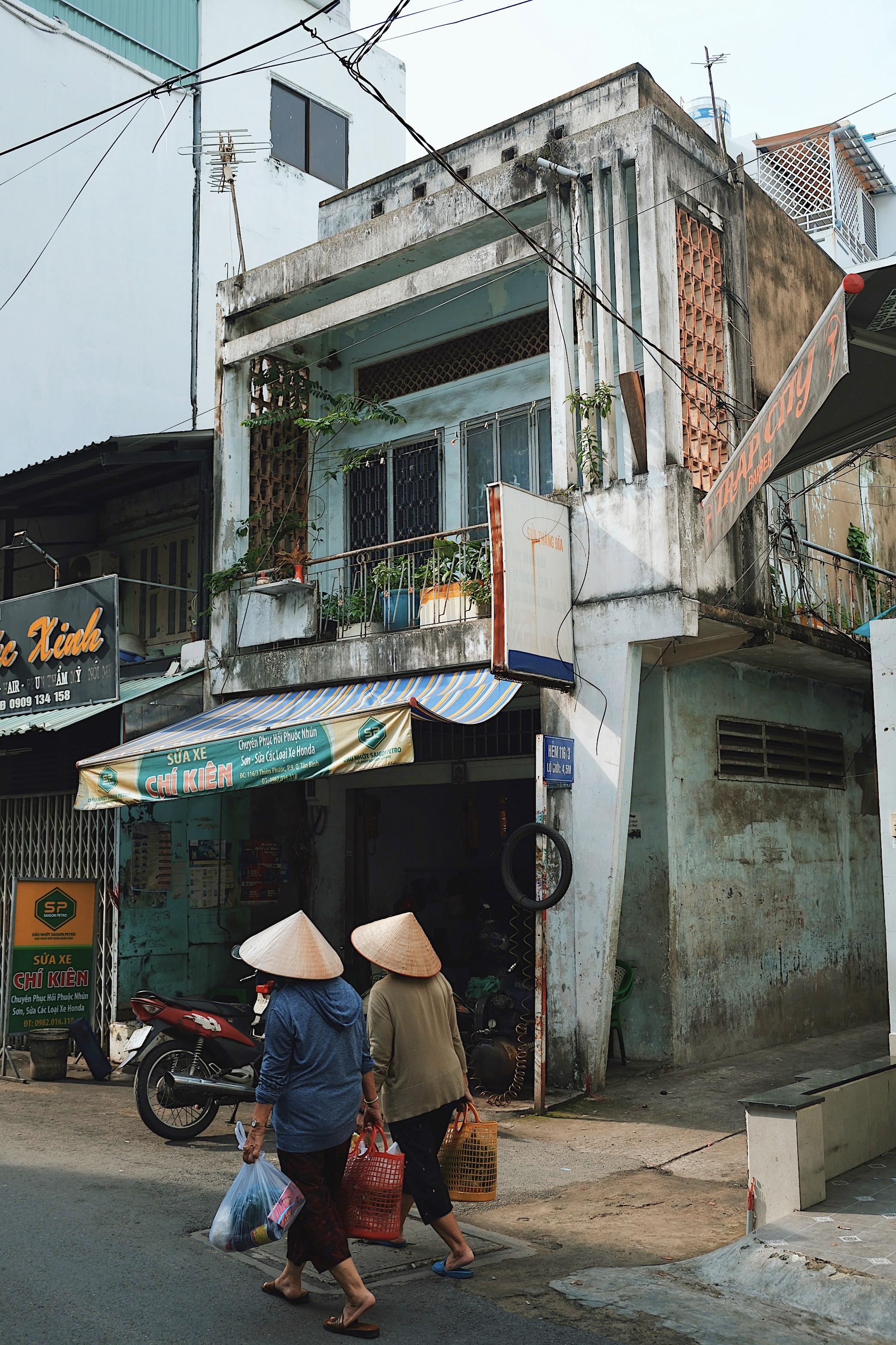 Everyday life in Ho Chi Minh City, Vietnam: Two women wearing conical hats walking past modernist shophouses with tropical design elements.