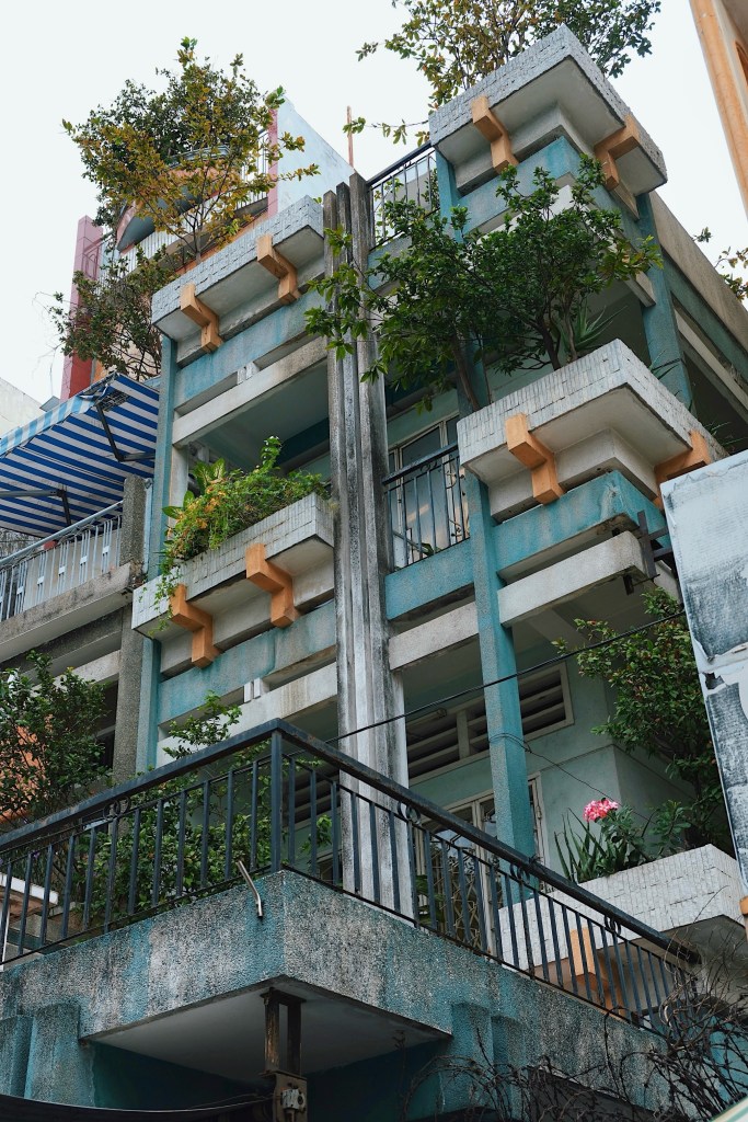 A contemporary modernist shophouse in Ho Chi Minh City, featuring a striking concrete vertical brise soleil complemented by lush, greenery-filled balconies that provide natural shade.