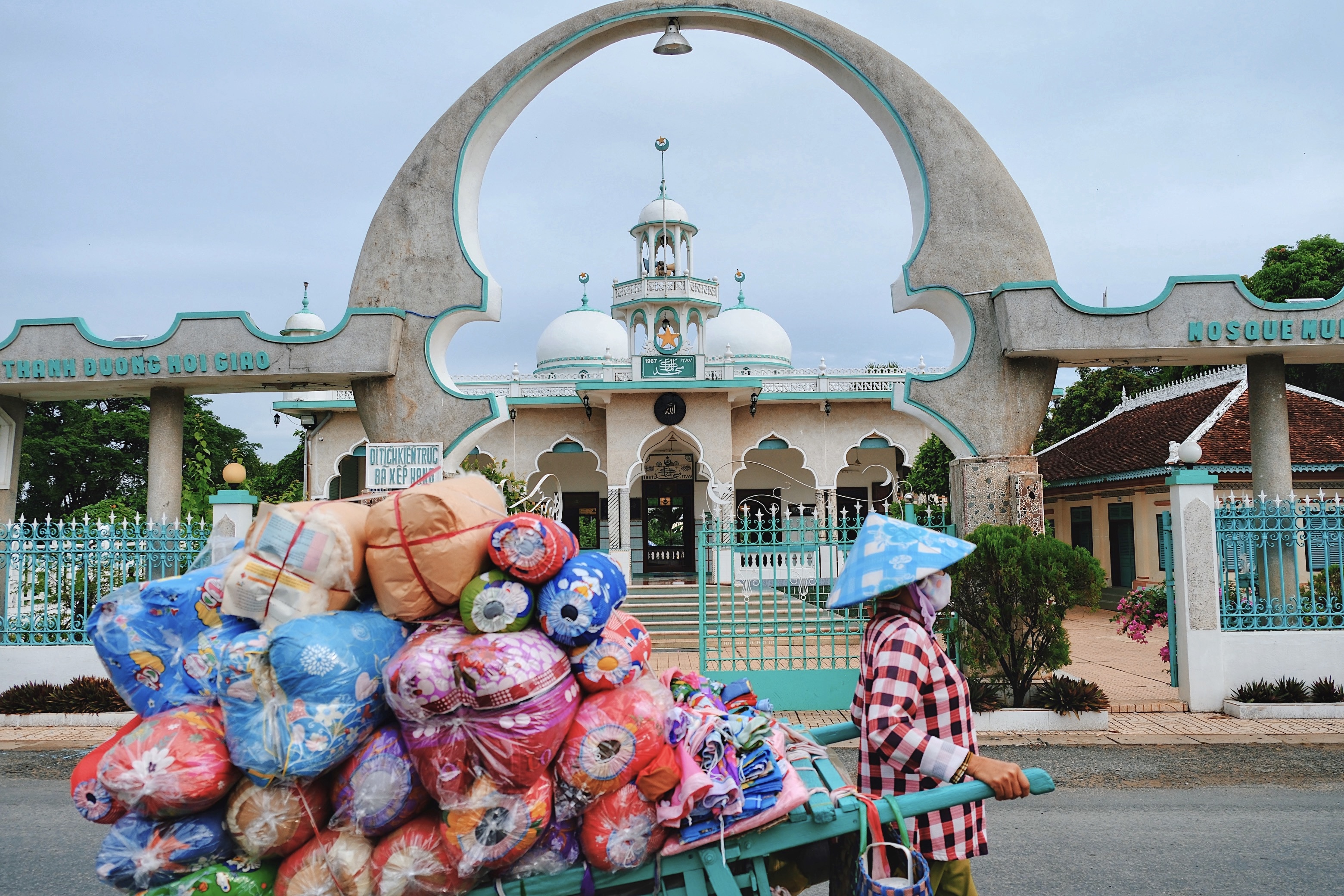 A bustling market street in Vietnam where a woman in a traditional conical hat pushes a cart loaded with colorful textiles past an ornate concrete mosque entrance