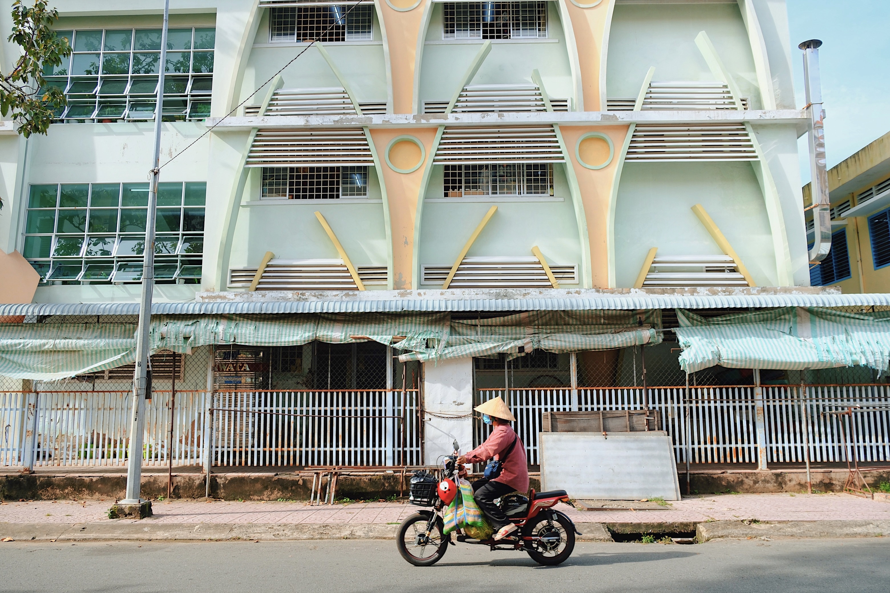 A pale green mid-century modern school building in Vietnam with curved concrete features, while a motorbike rider in a conical hat drives by