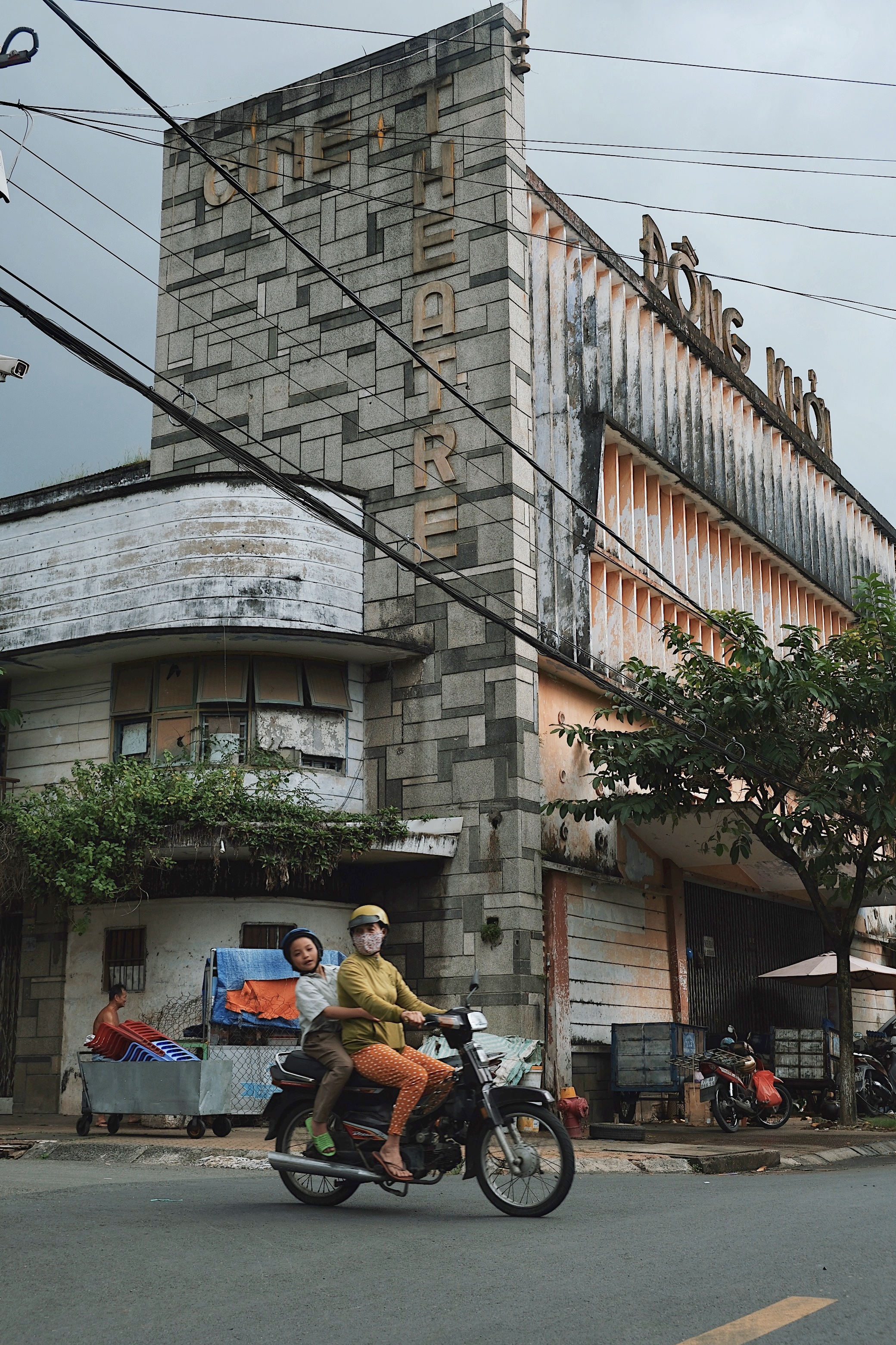 A weathered mid-century modern theater in Vietnam (Ben Tre) with a Brutalist-style concrete facade, as a woman and child ride past on a motorbike