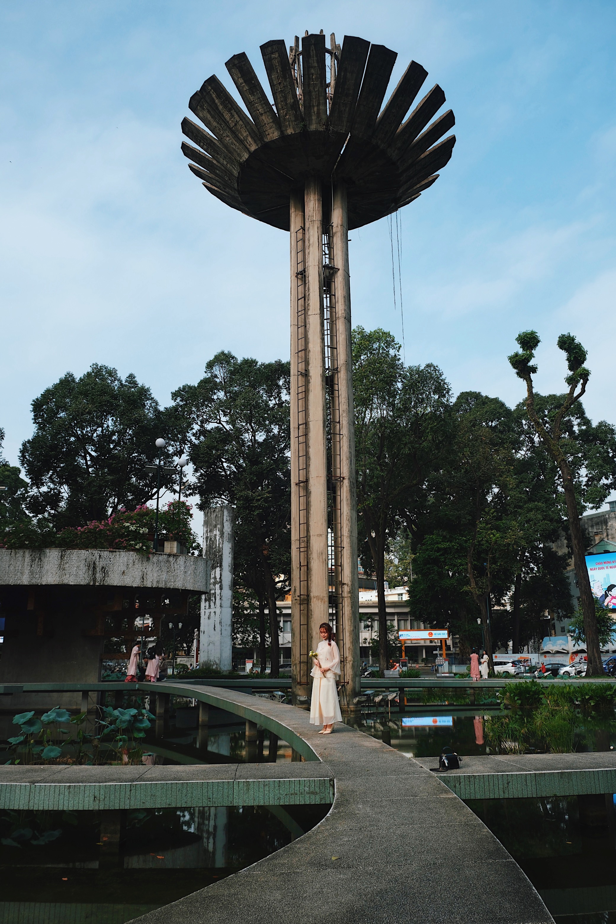  A concrete towering lotus-shaped Brutalist monument in Ho Chi Minh Vietnam, with a reflective pond and a woman in a flowing white dress standing on a curved bridge