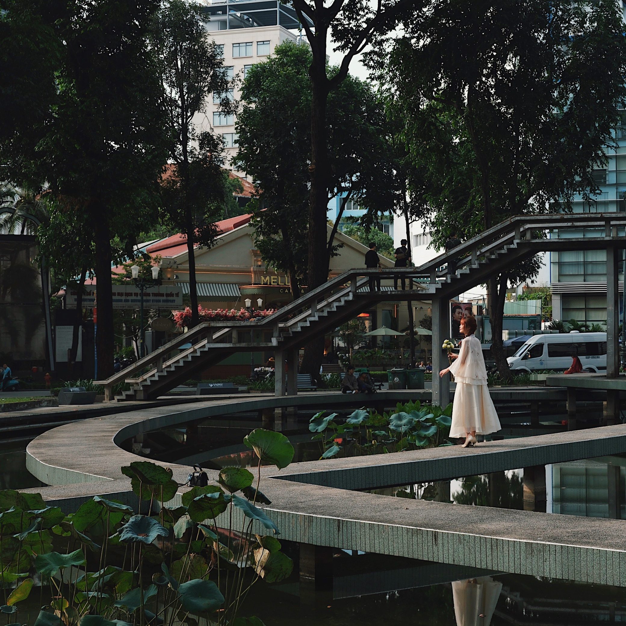 A peaceful urban park in Ho Chi Minh Vietnam featuring modernist pedestrian bridges over a pond, with a woman in a traditional outfit posing