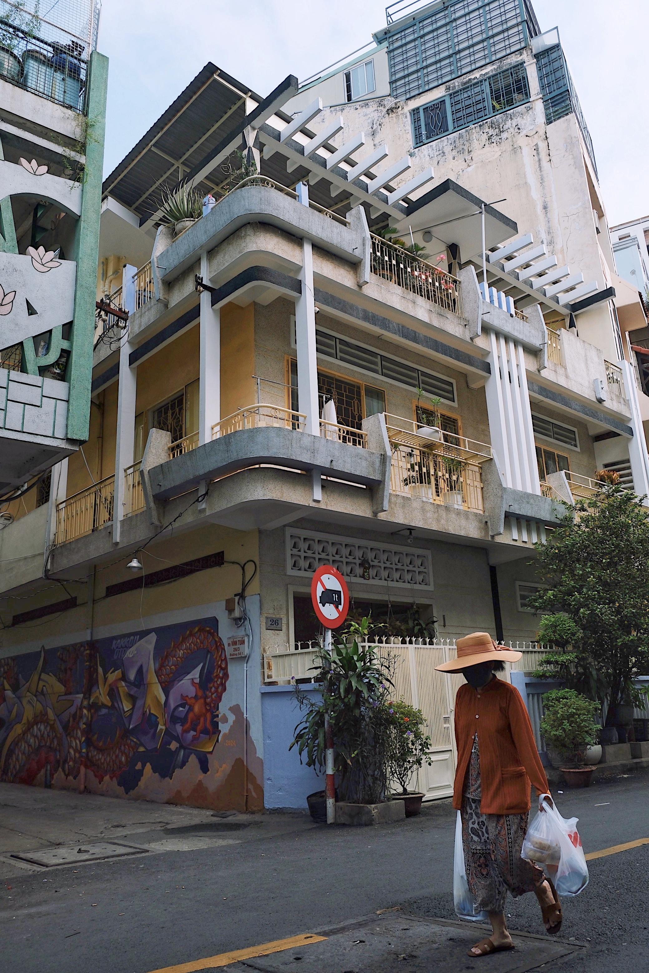 A woman wearing a conical hat shops for groceries in Ho Chi Minh City, standing in front of a striking modernist building with geometric balconies and urban textures.