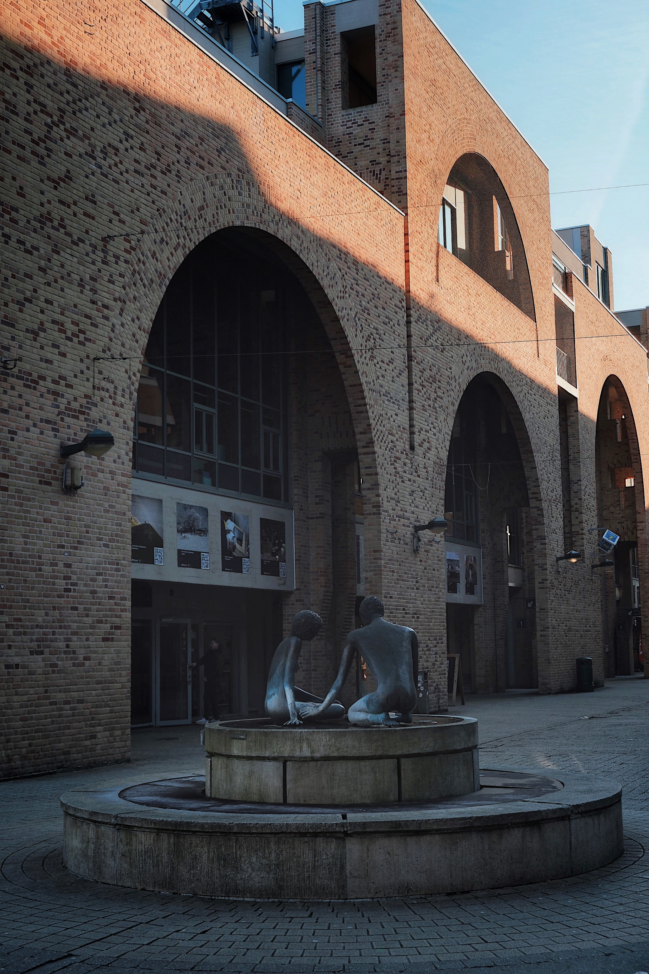 A circular stone fountain with abstract human sculptures in Louvain-la-Neuve’s university square, framed by arched brick architecture and soft golden light.