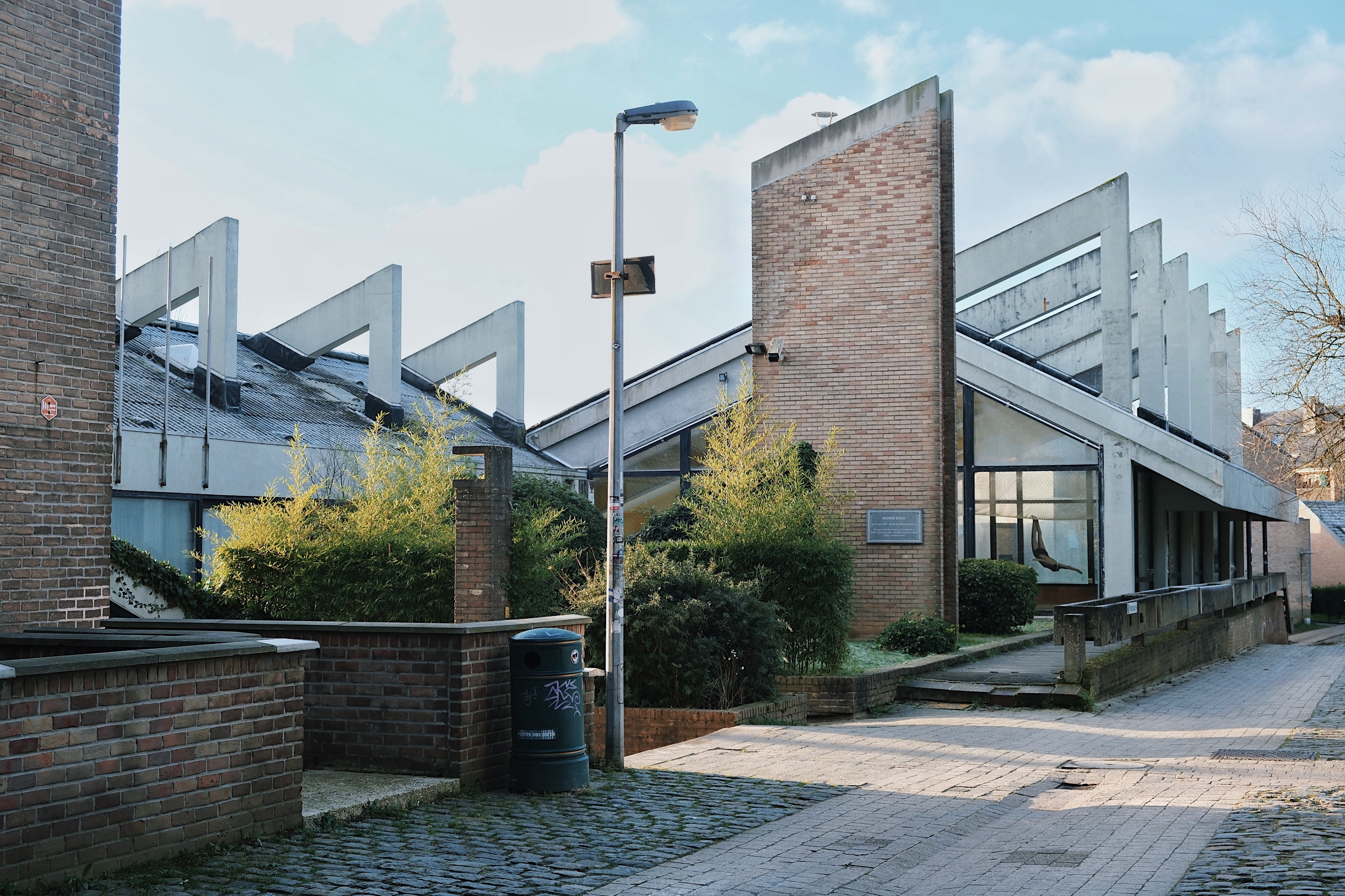 A striking Brutalist swimming pool in Louvain-la-Neuve, featuring angular concrete structures, large glass windows, and a red brick accent wall.