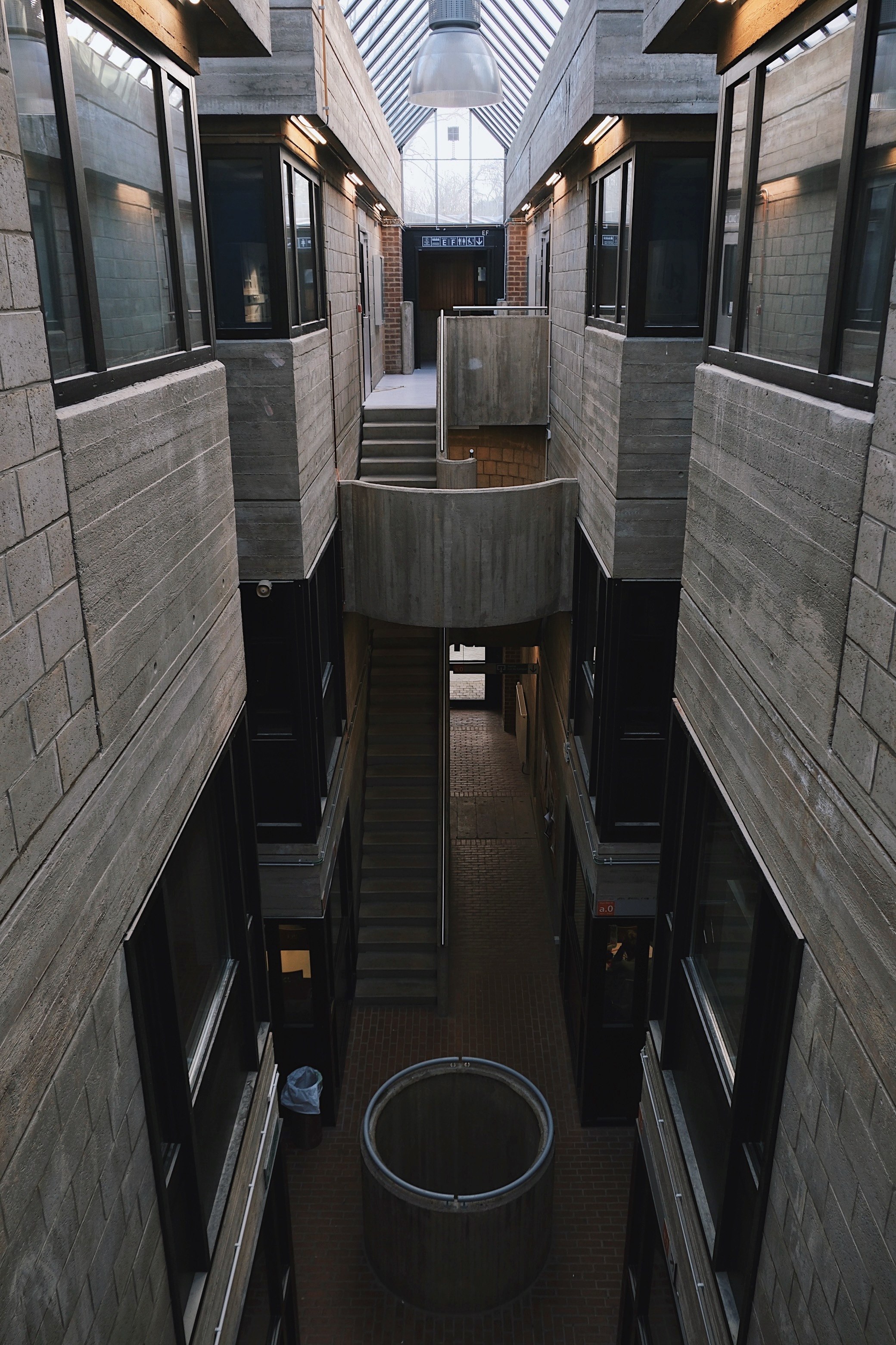 A Brutalist academic corridor in Louvain-la-Neuve’s Pierre Curie Building, featuring exposed concrete walls, skylights, and an industrial staircase leading to upper floors.