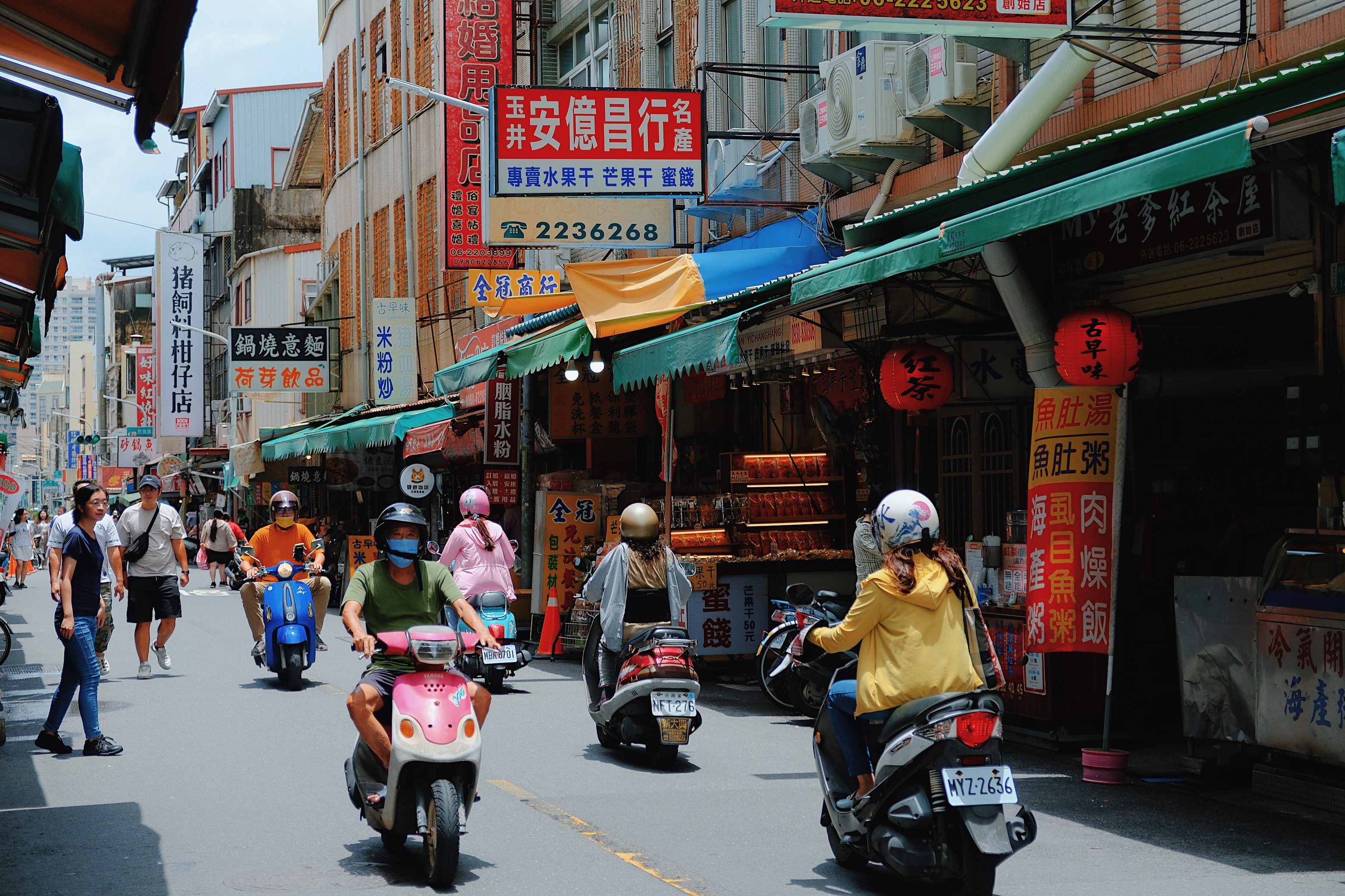 A bustling street market in Tainan, Taiwan, where vendors sell goods under colorful awnings as scooters navigate the narrow, sign-filled road.