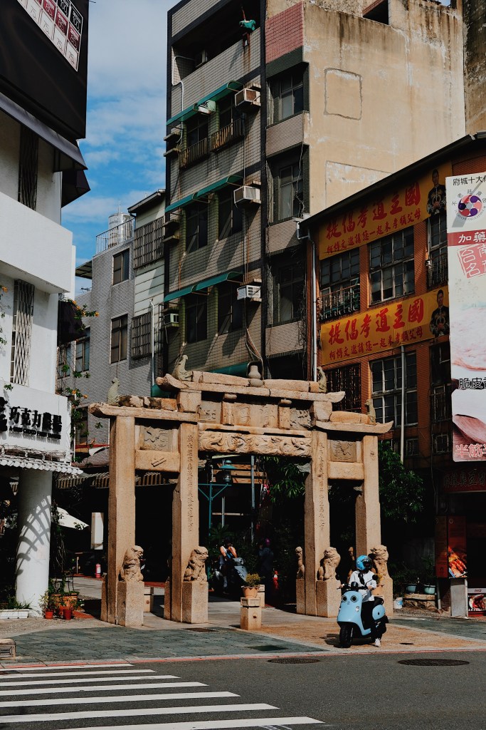  A traditional stone gateway in Tainan, Taiwan, standing amid modernist buildings and colorful signage, representing the city's layered architectural history.
