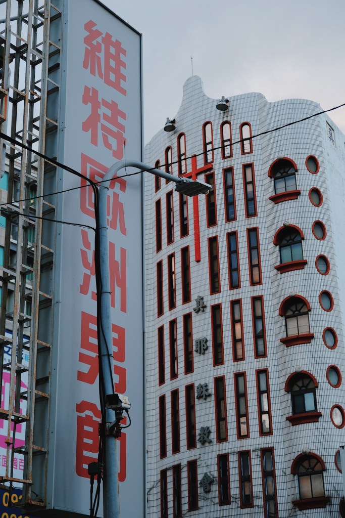 A curved, white-tiled modernist church in Tainan, Taiwan, adorned with red accents and an arched cross motif, reflecting Japanese architectural influences.