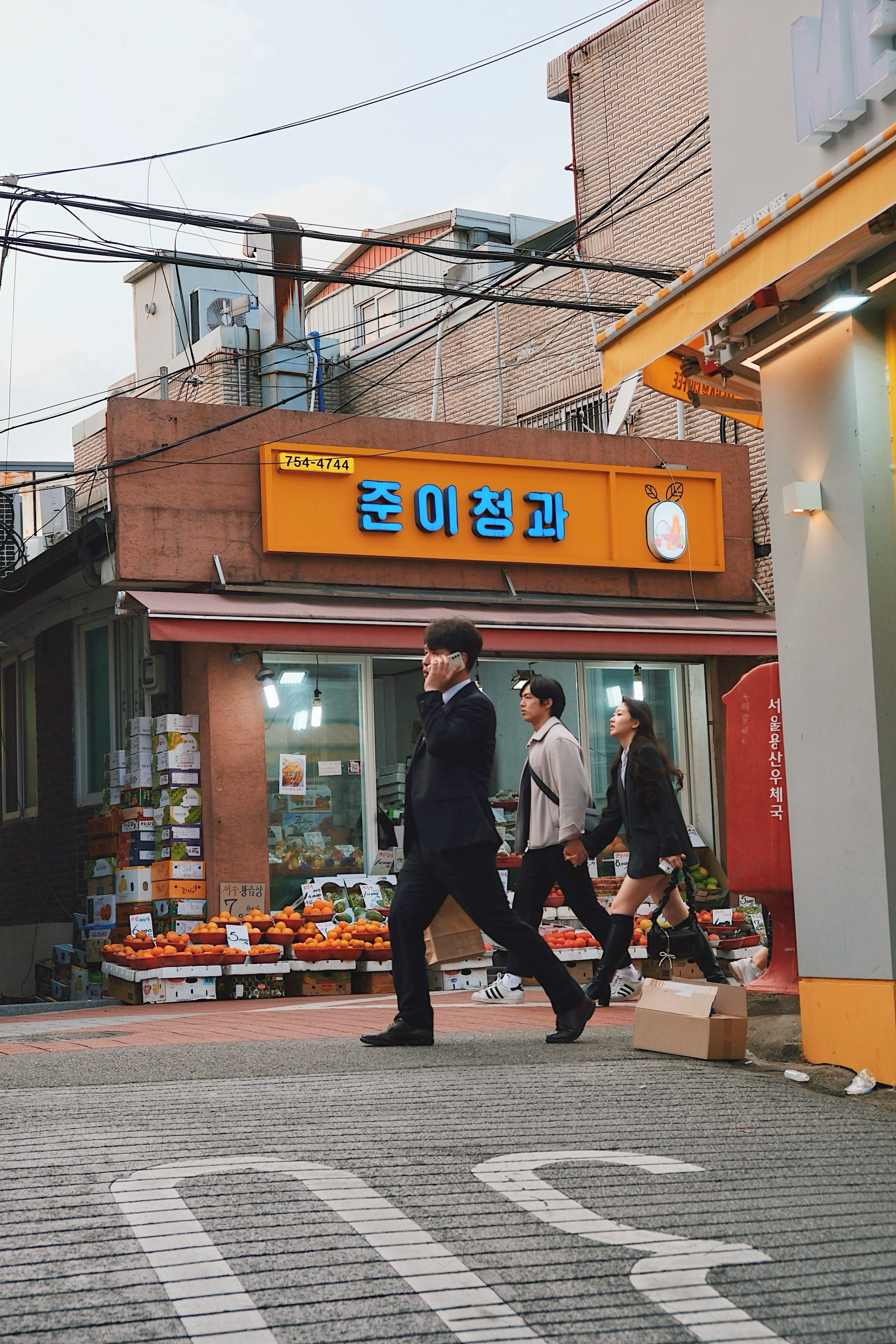 Street scene in Haebangchon, Seoul, showing a compact urban corner store beneath dense electrical lines and vibrant signage, contrasting human motion against static architecture