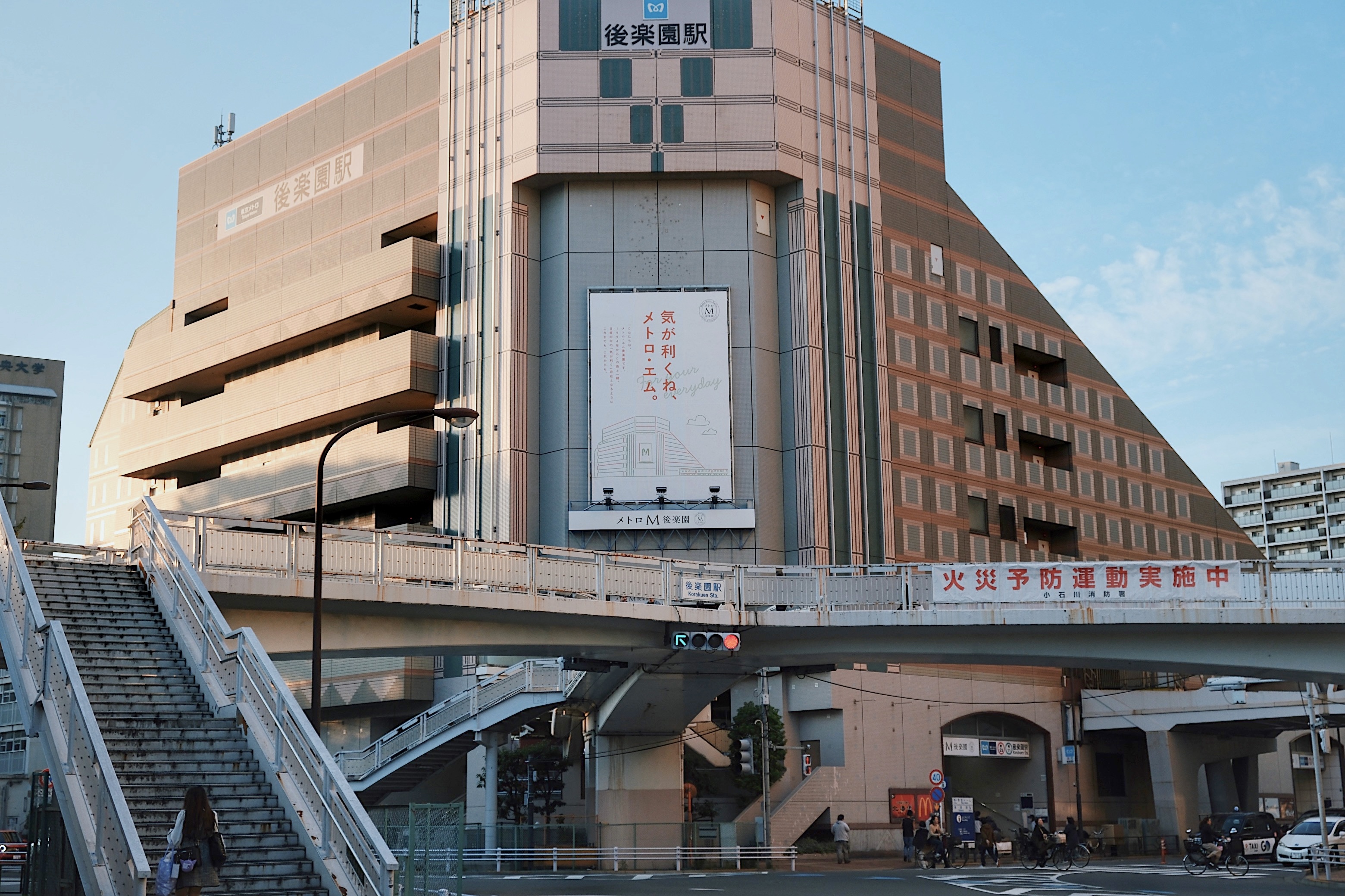 The modernist, tiered structure of Korakuen Station in Tokyo, with an elevated pedestrian walkway and a grand beige facade.