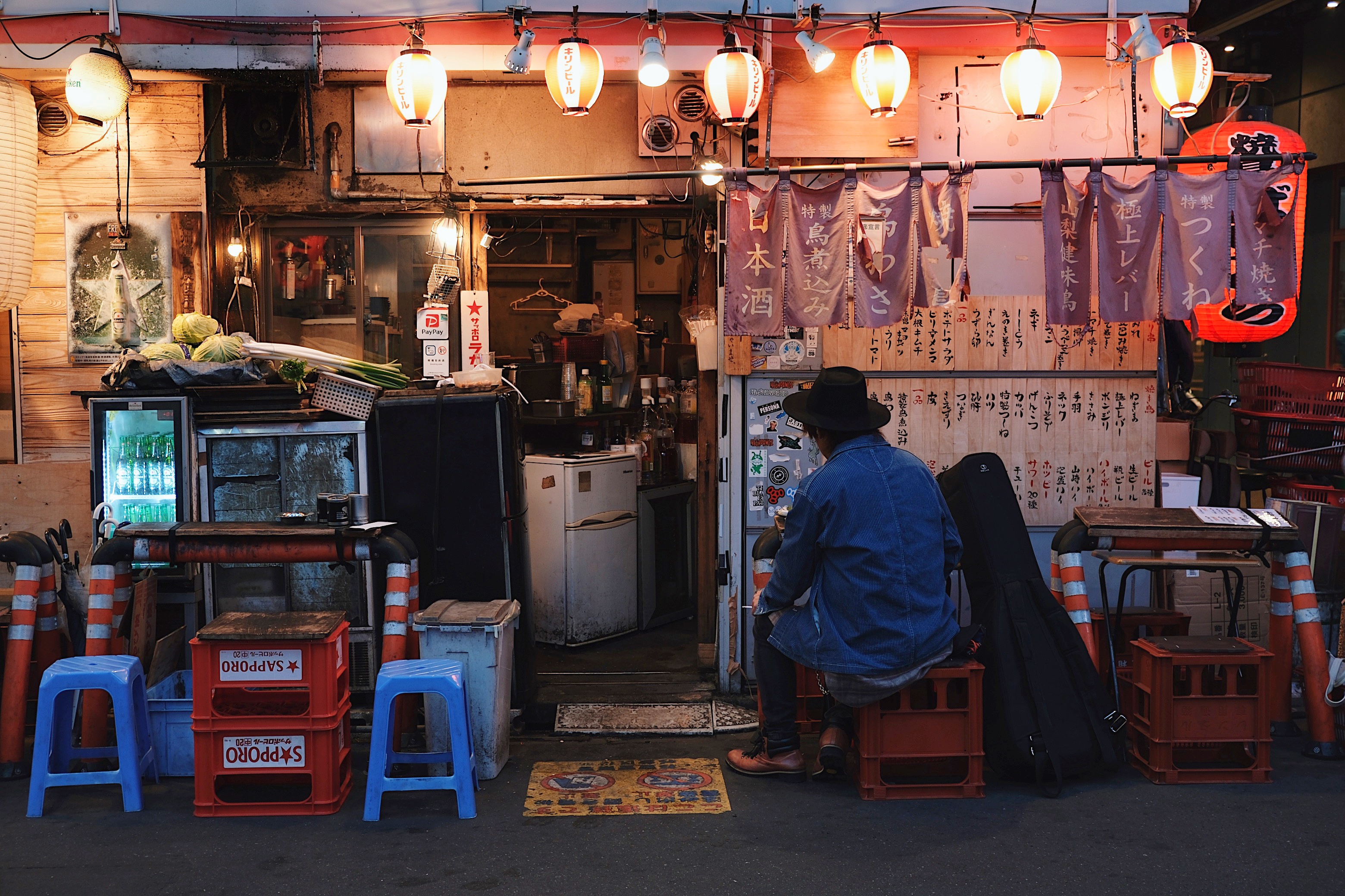 A rustic, hole-in-the-wall izakaya in Koenji, where a man in a denim jacket and hat sits alone under the soft glow of red paper lanterns.

