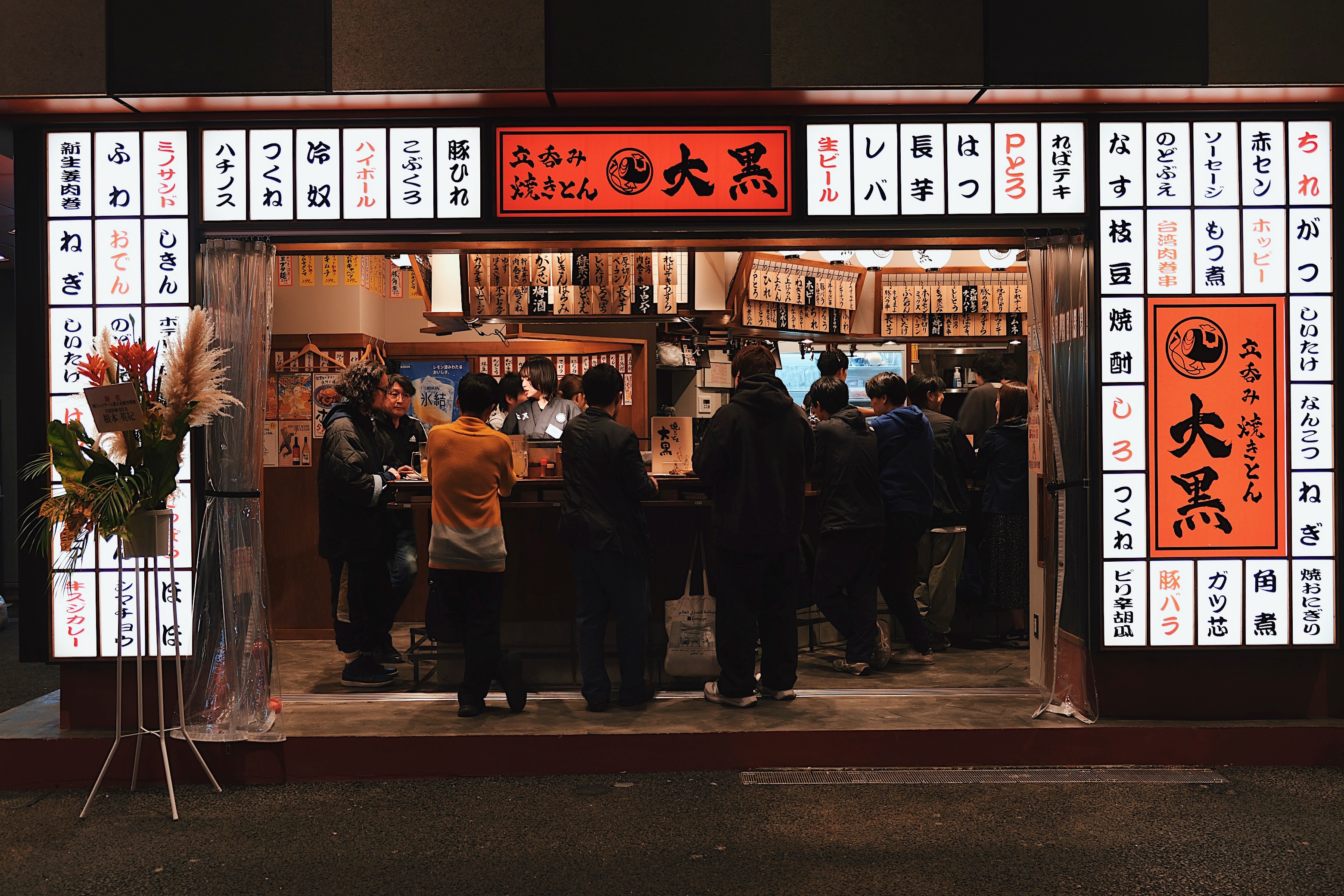 A stylish izakaya in Koenji, Tokyo, where patrons enjoy drinks under glowing lanterns and a facade covered in traditional Japanese calligraphy.