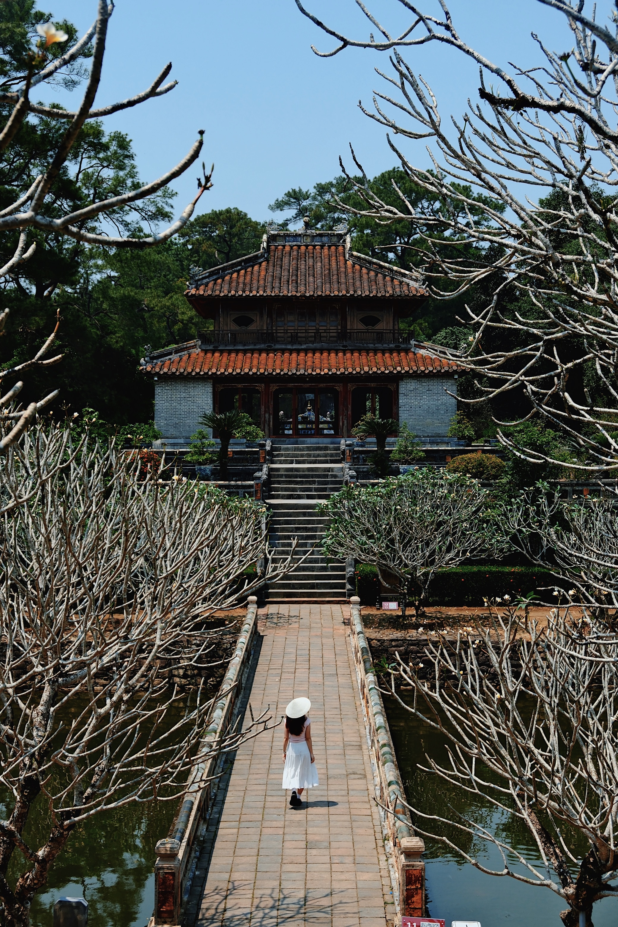 A woman in a traditional white áo dài walks across a wooden bridge leading to the Thien Mu Pagoda, a historic Buddhist temple in Hue, surrounded by lush greenery and a tranquil pond.