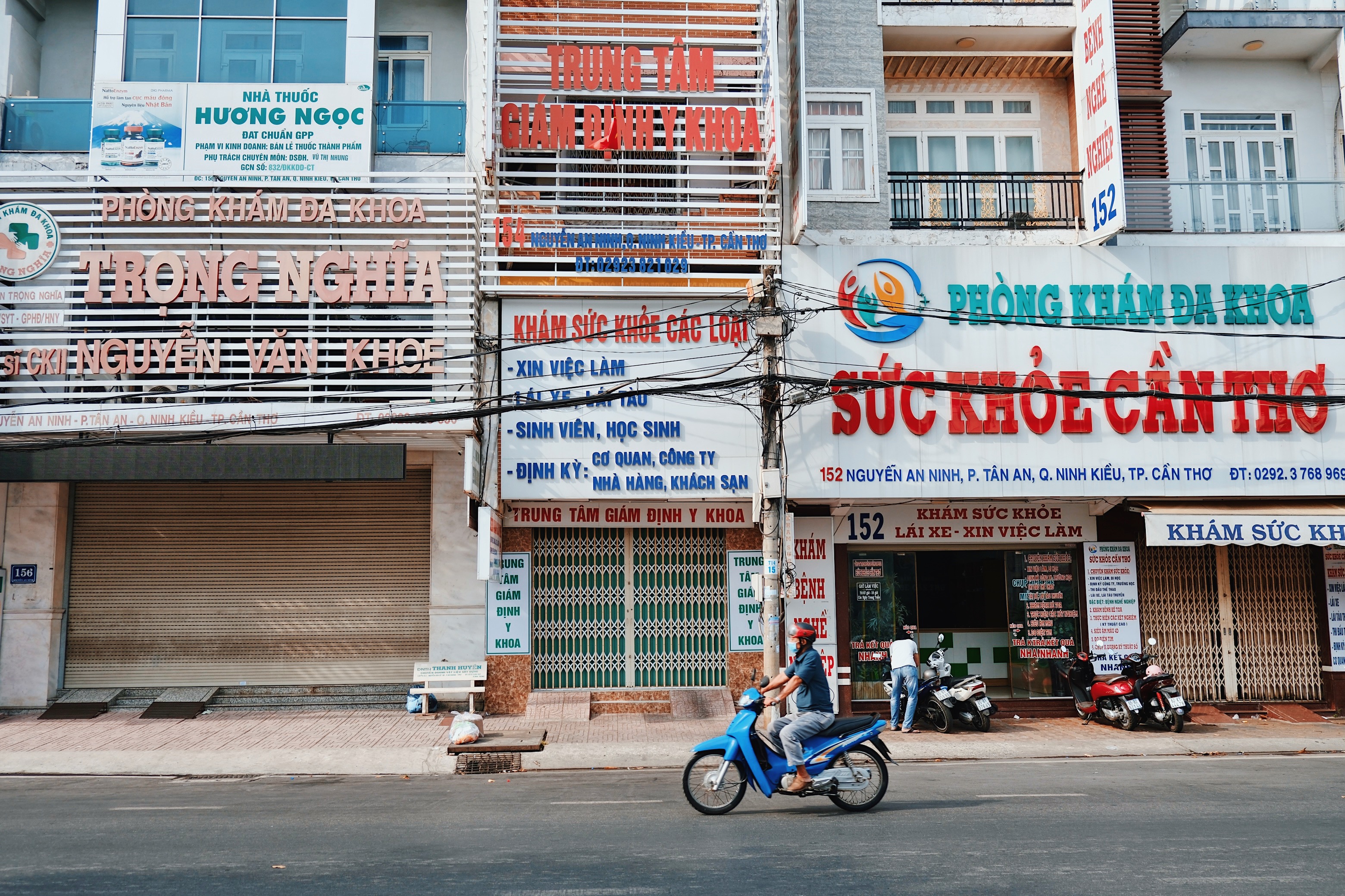 A street in Cần Thơ, Vietnam, lined with medical clinics and businesses, where a motorbike rider passes by colorful, overlapping signage.
