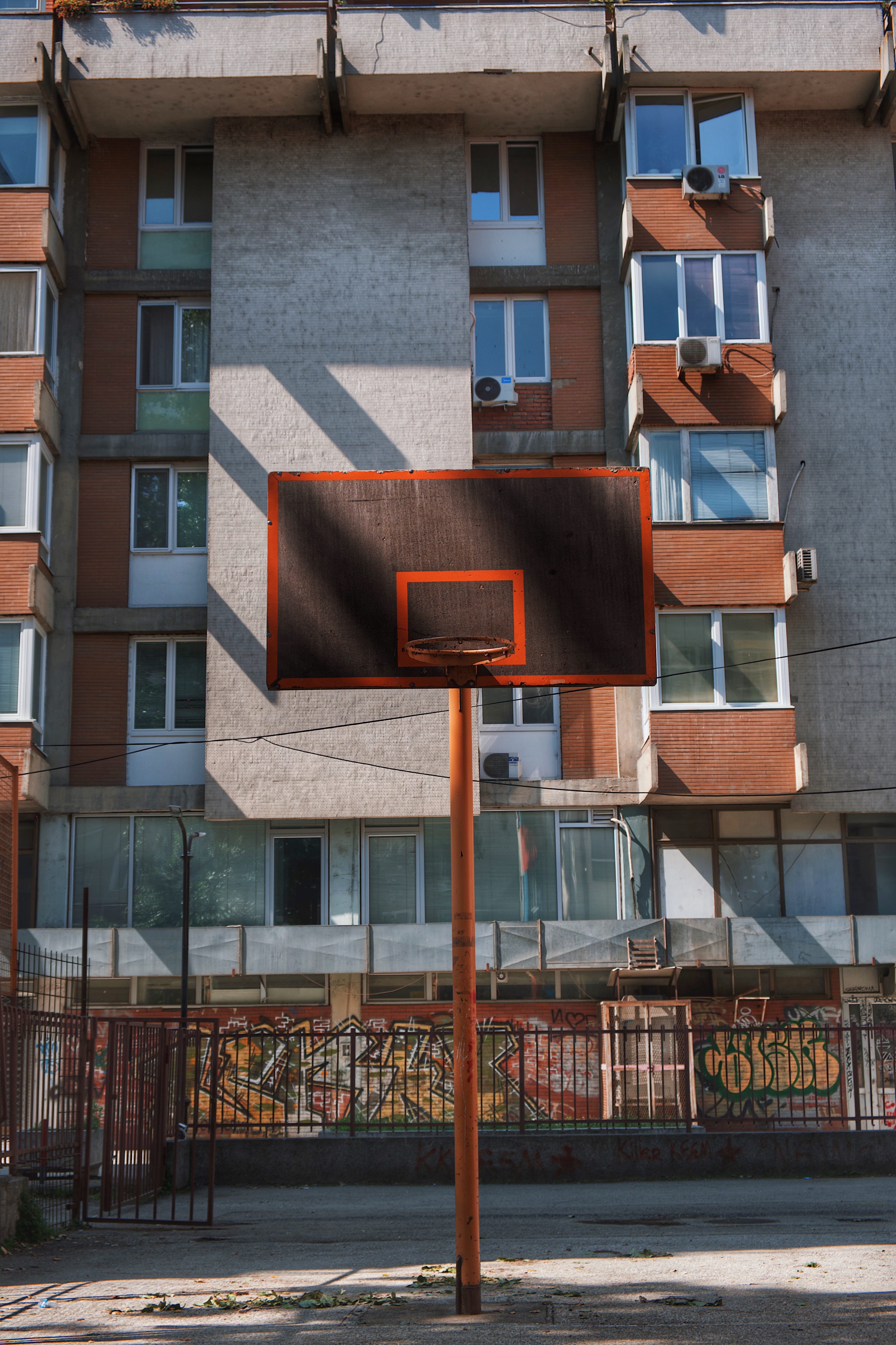 Outdoor basketball hoop in a residential area near Maksim Gorki, Skopje, surrounded by socialist-era apartment blocks and graffiti-covered fencing