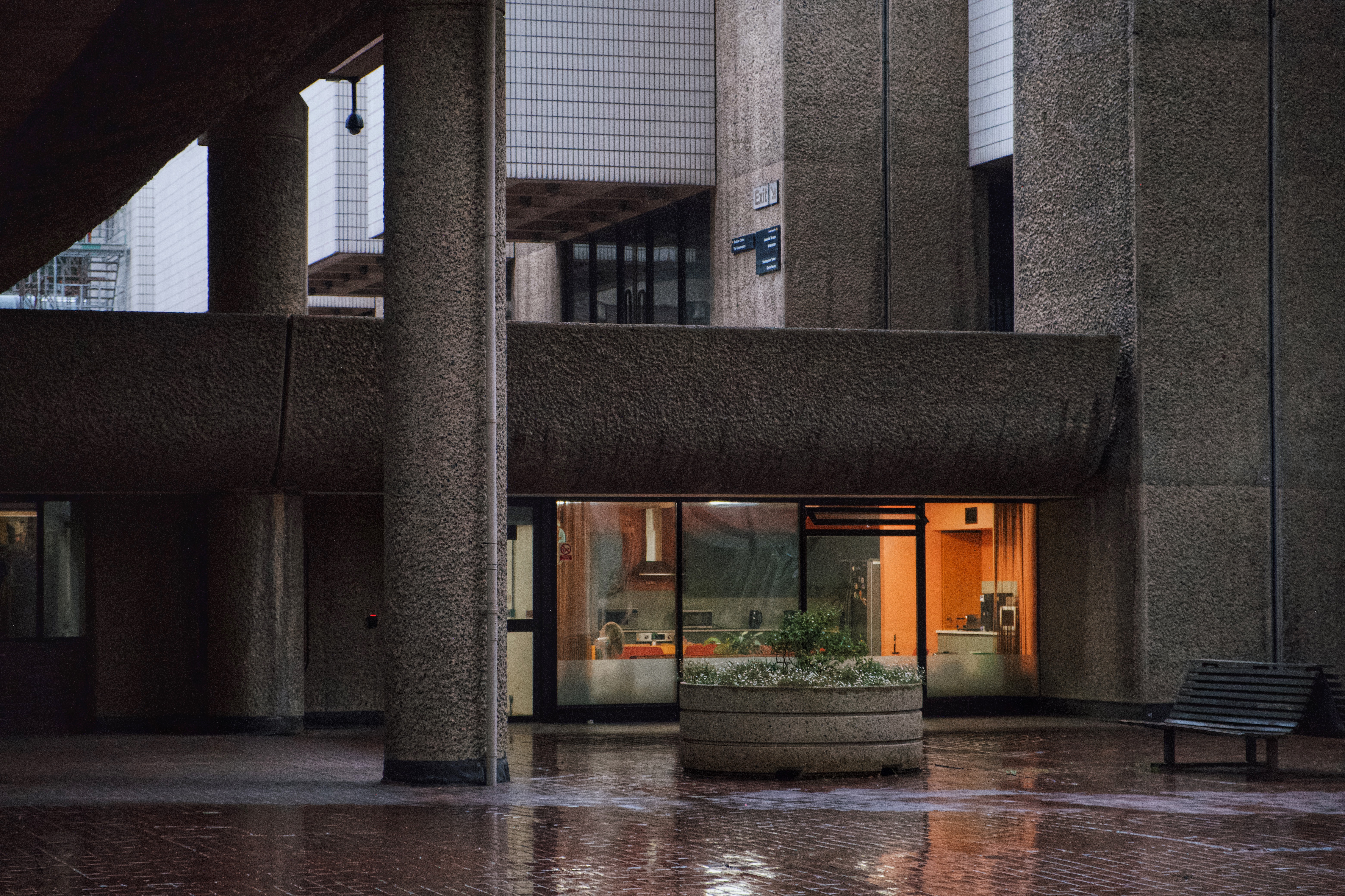 Brutalist courtyard at the Barbican Estate in London, with heavy concrete columns, soft reflections on wet brick pavement, and a glimpse of warm interior light