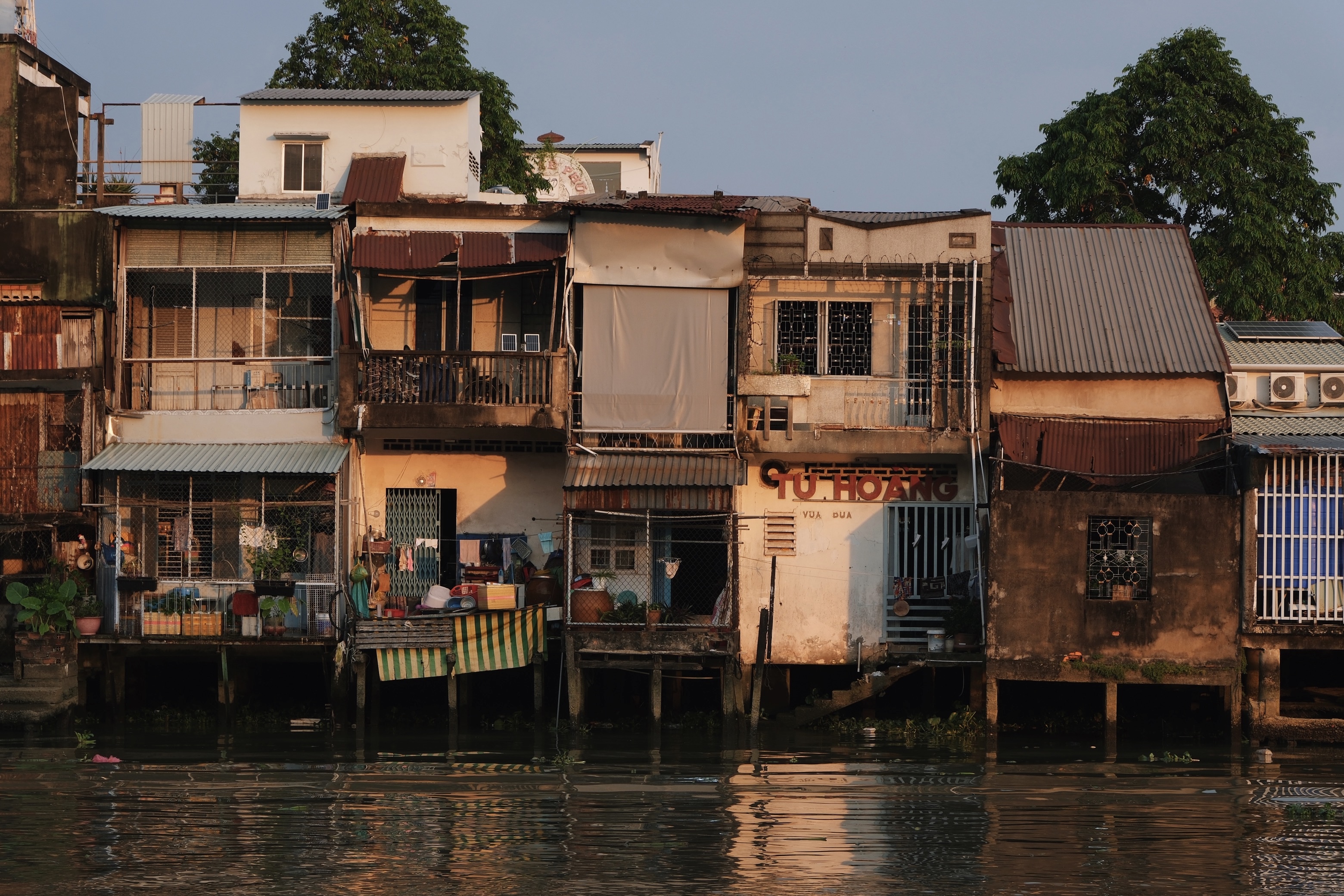 Riverside scene in My Tho, Mekong Delta, featuring vernacular stilt houses and modernist architecture reflected in the water among colorful shopfronts and signage.