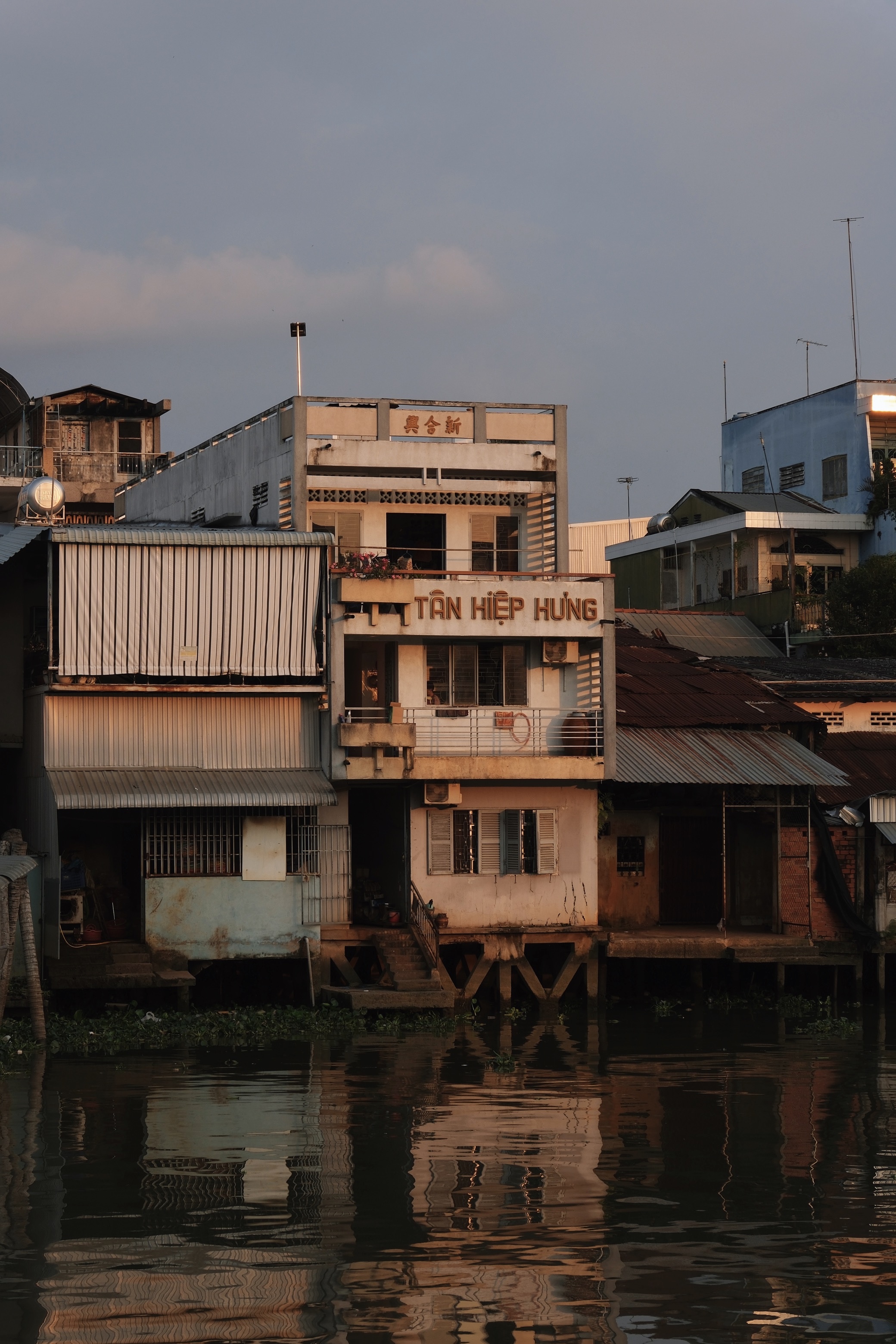 Waterfront architecture in My Tho, Mekong Delta, at sunset, with stilt houses and modernist buildings reflected in the river alongside traditional Asian shopfronts and signage.