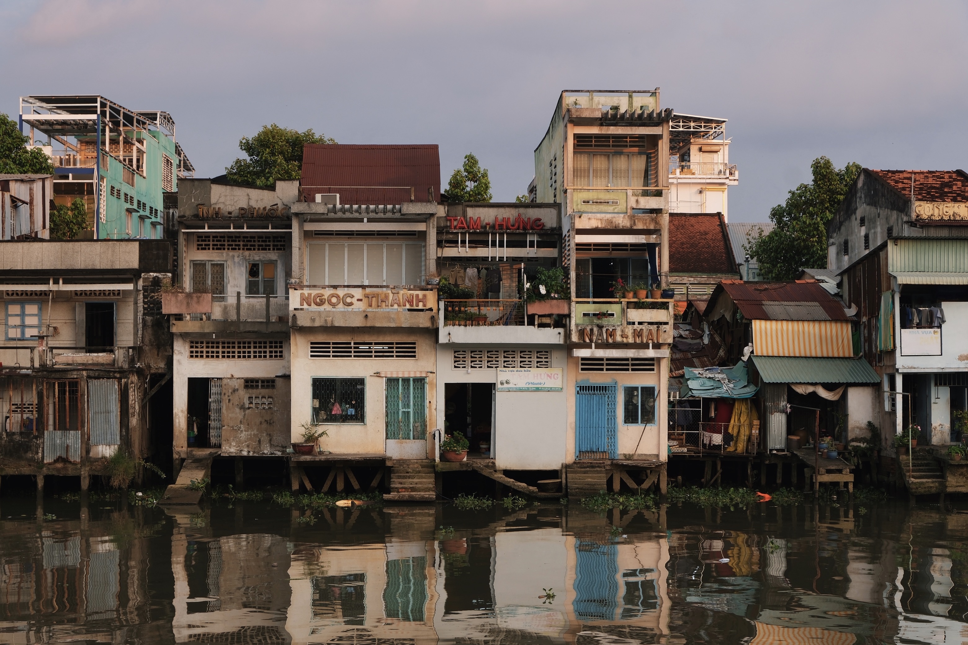 Sunset view of My Tho town in Vietnam’s Mekong Delta, showing stilt house architecture and mid-century modernist façades mirrored in the calm waterfront.